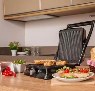 Oster GRL10 aberta, vista de lado em cima de bancada de cozinha, com bifes de frango no grill.